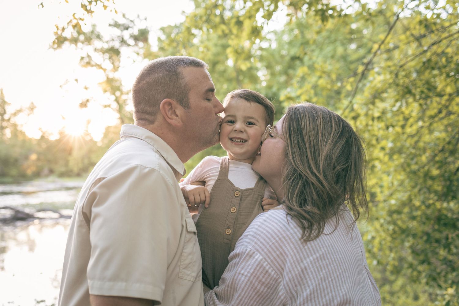 Parents kissing their smiling toddler on the cheeks outdoors in golden hour sunlight near a tree-lined riverbank.