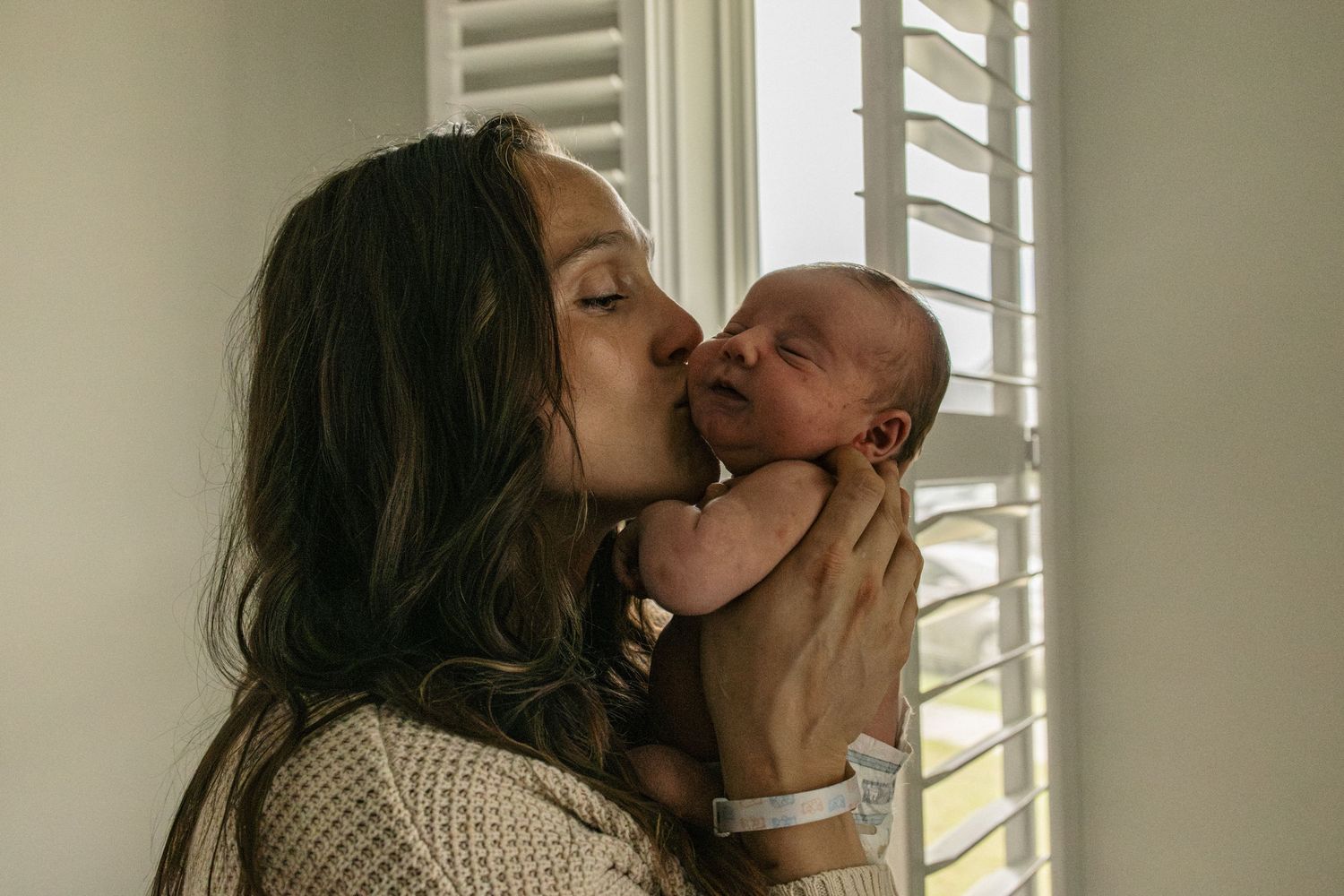 A tender moment between parent and newborn baby by a window with white shutters.
