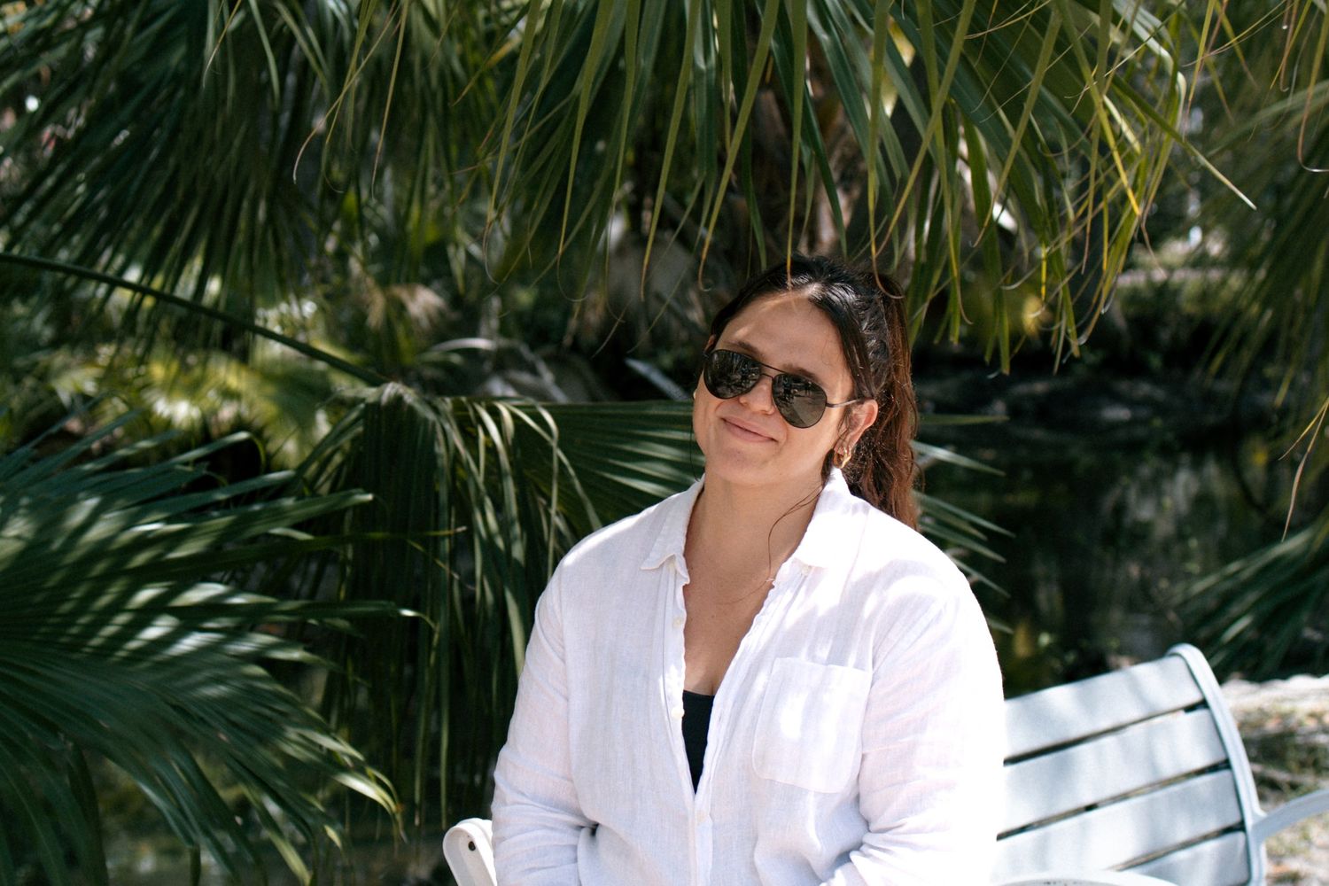 Someone wearing a white button-down shirt and sunglasses sits outdoors among tropical palm trees on a sunny day.