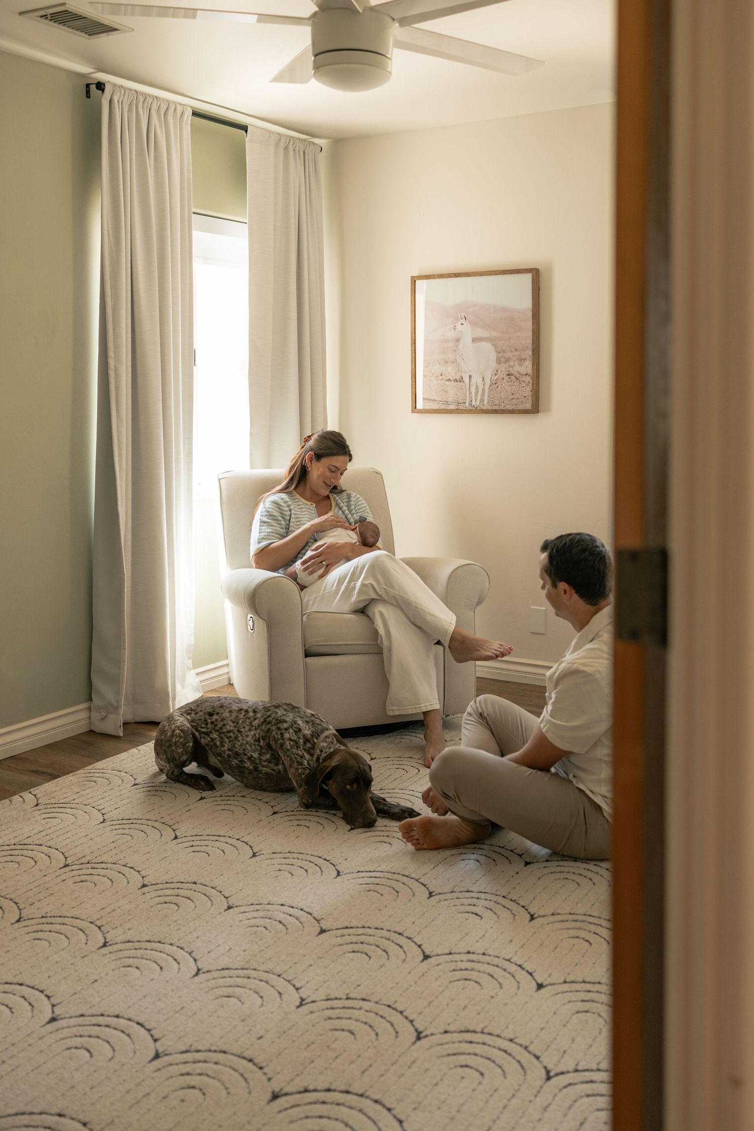 A mother nurses her newborn in a nursery glider while her partner sits on the floor beside her and their dog rests nearby.