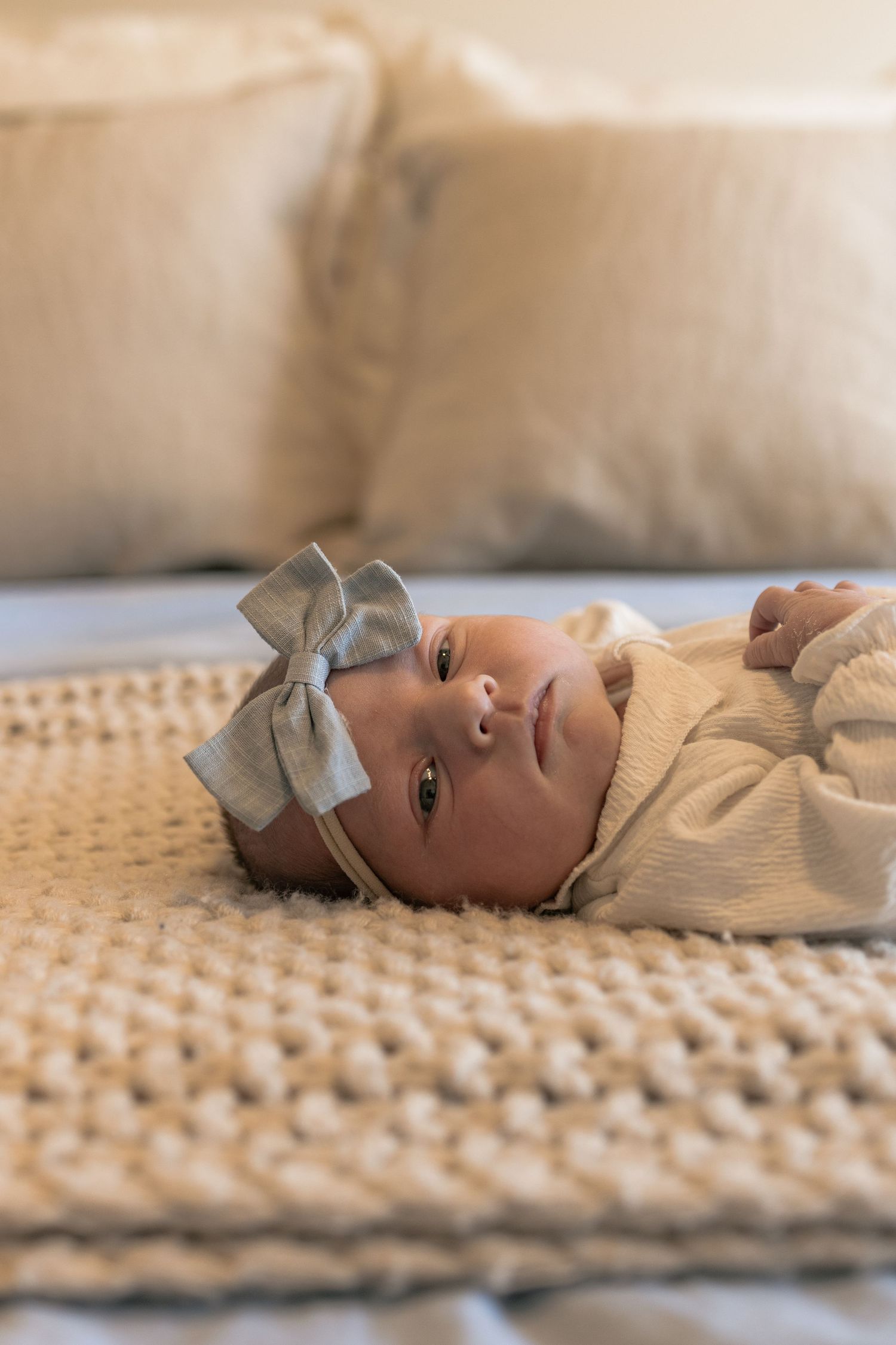 Newborn baby girl lying on chunky knit blanket wearing cream outfit and gray bow headband.