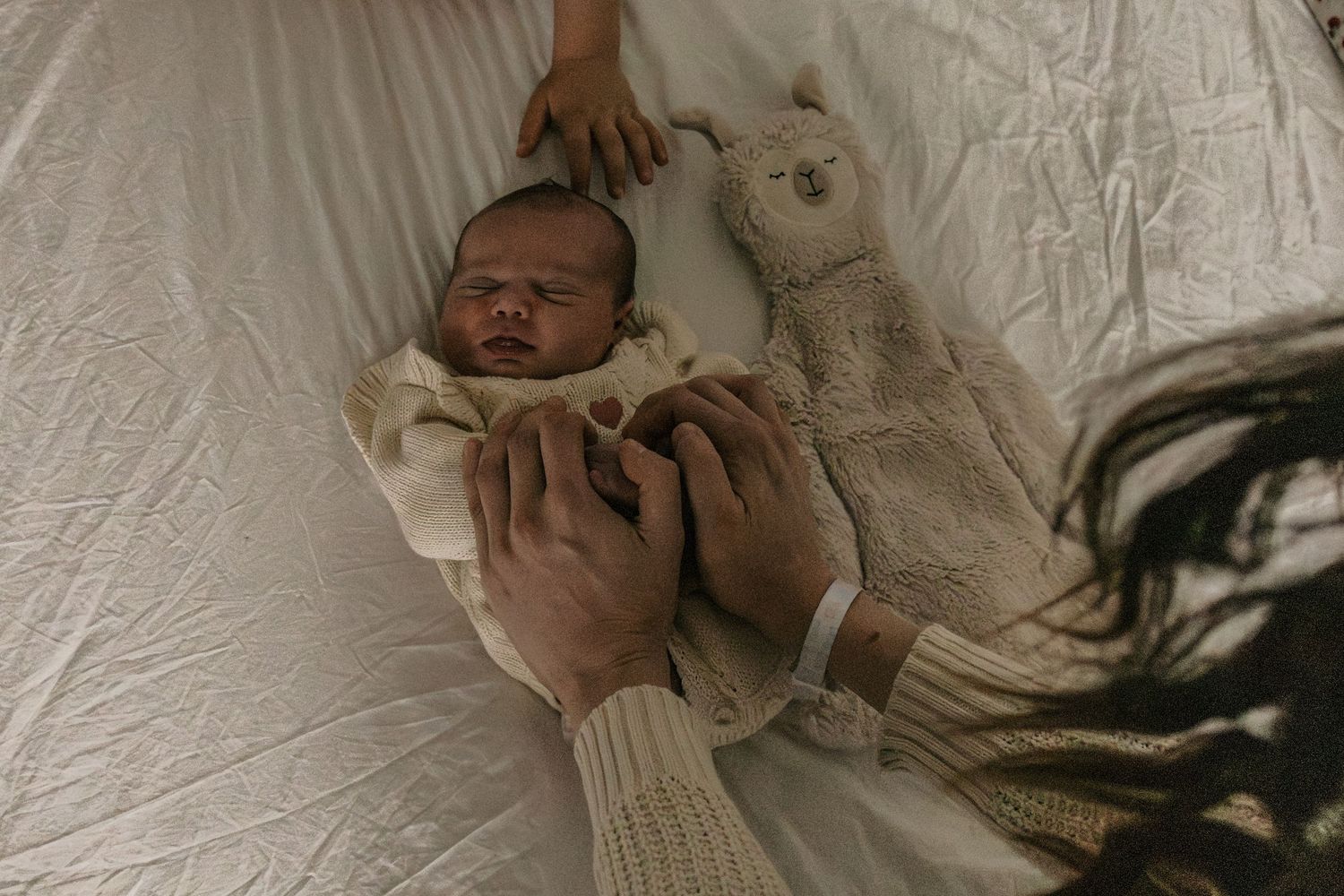 A newborn baby lies on white bedding while being gently held by hands in a knit sweater.