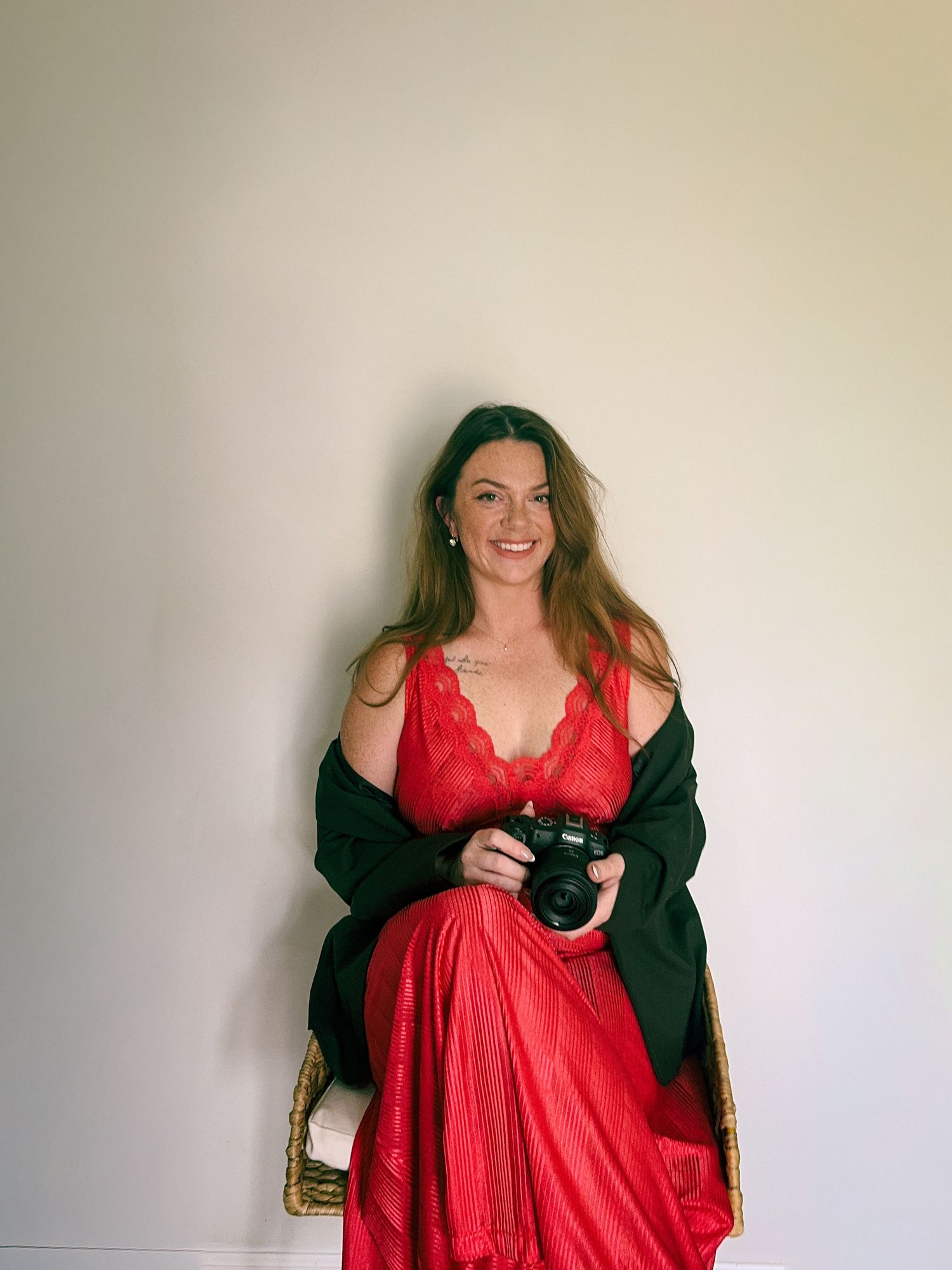 A person in a flowing red dress and black cardigan poses against a light-colored wall.