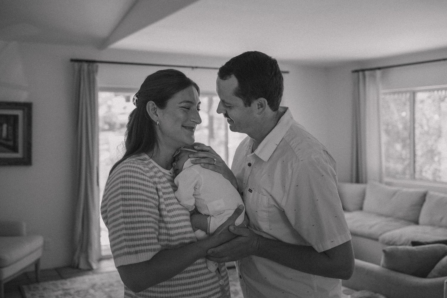 Black and white photo of a couple smiling at each other while holding their newborn baby in a cozy living room.