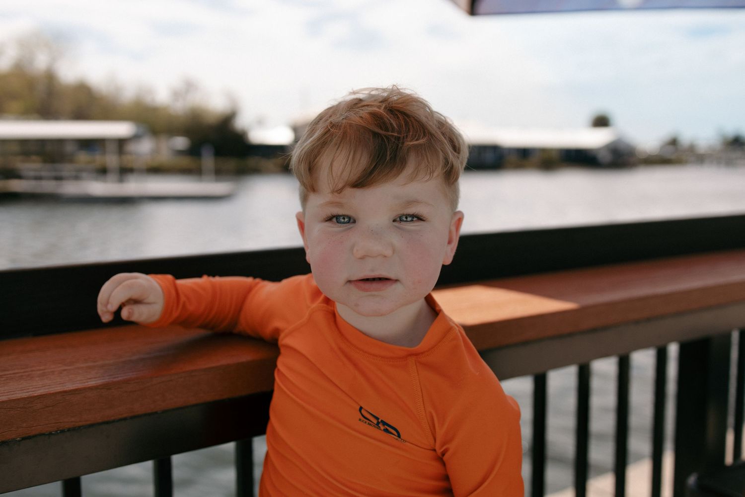 A young child wearing an orange shirt sits on a wooden railing outdoors on a sunny day.