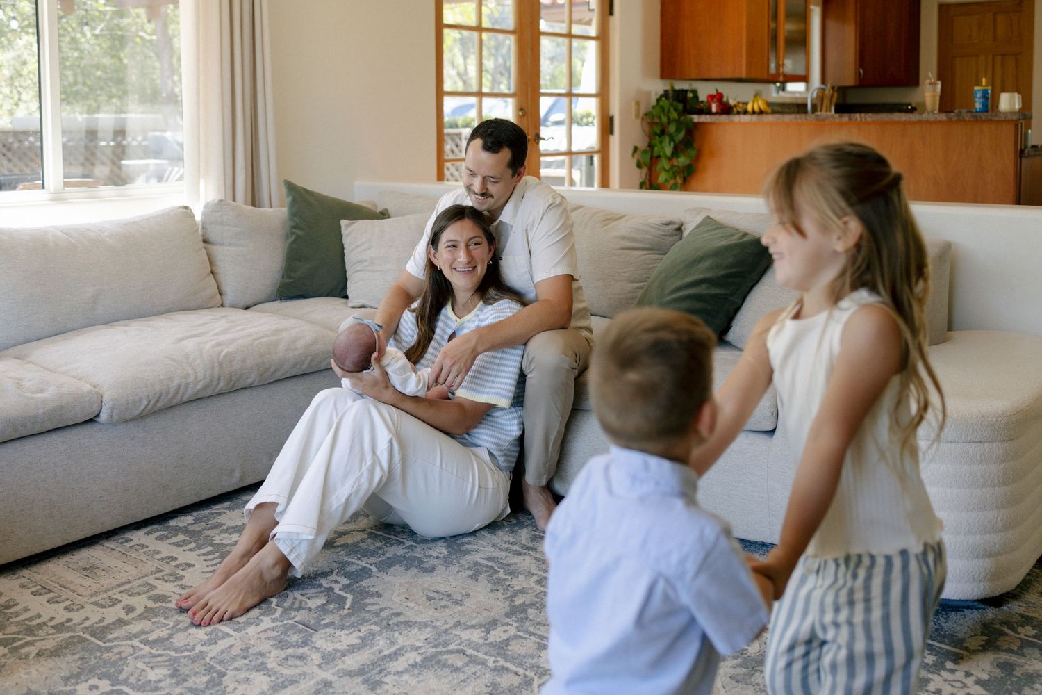 Family of five relaxing in a bright living room, parents sitting on floor with newborn while two children play nearby.