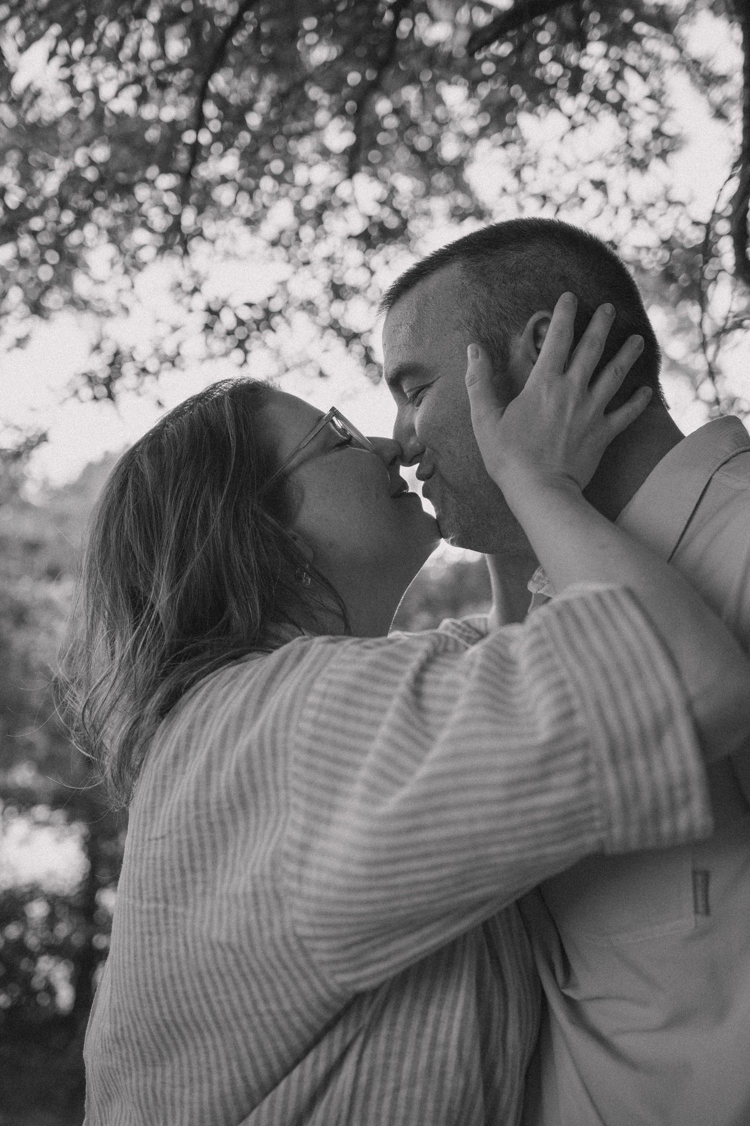 Black and white photo of a romantic couple sharing a tender kiss outdoors under a tree.