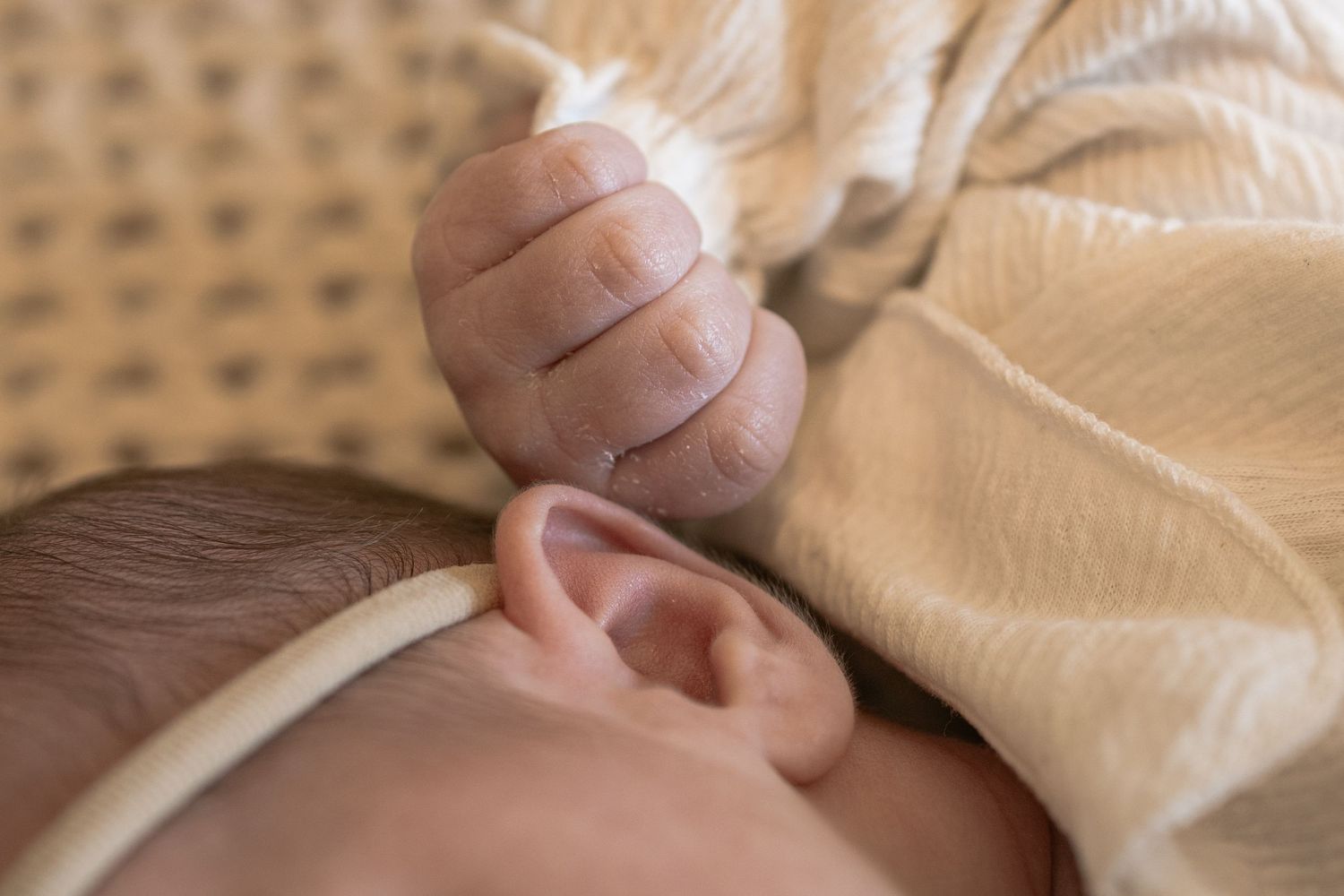 Close-up of sleeping newborn baby's tiny hand resting near ear wrapped in soft yellow blanket.