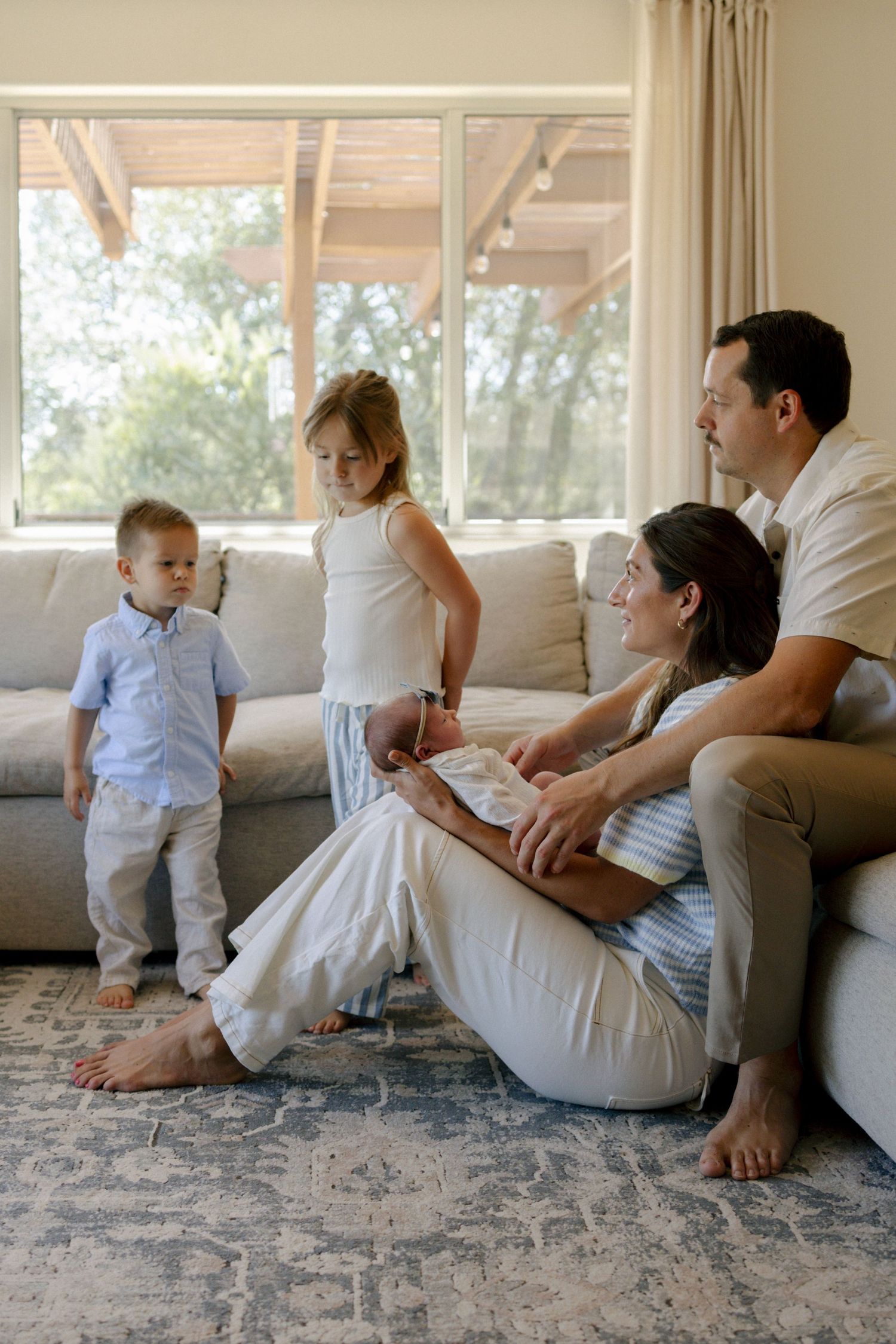 Family of four with newborn baby relaxing in bright living room, parents seated on floor while children stand nearby.