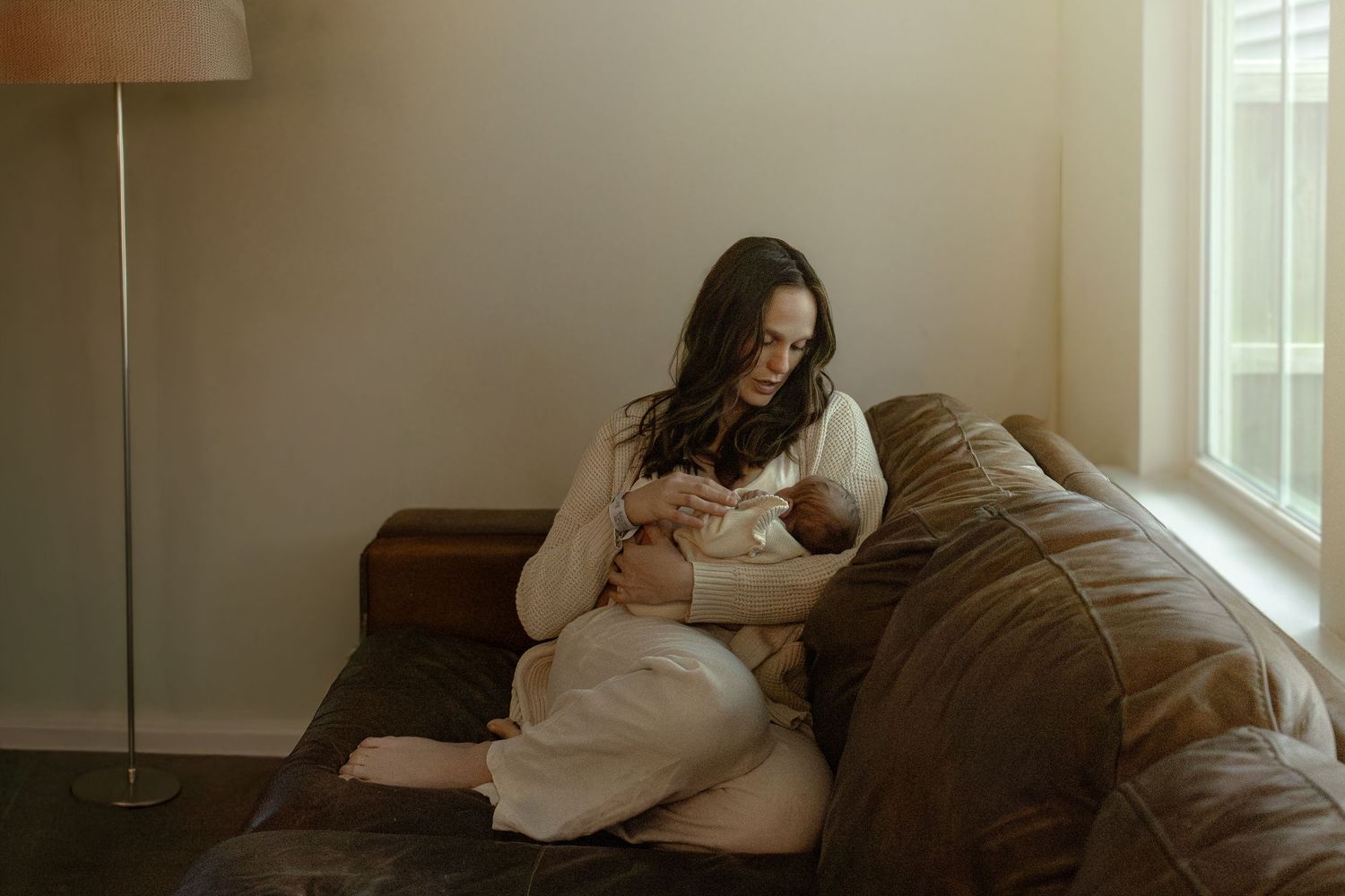 A person lies comfortably on a gray couch near a bright window in a cozy living room setting.