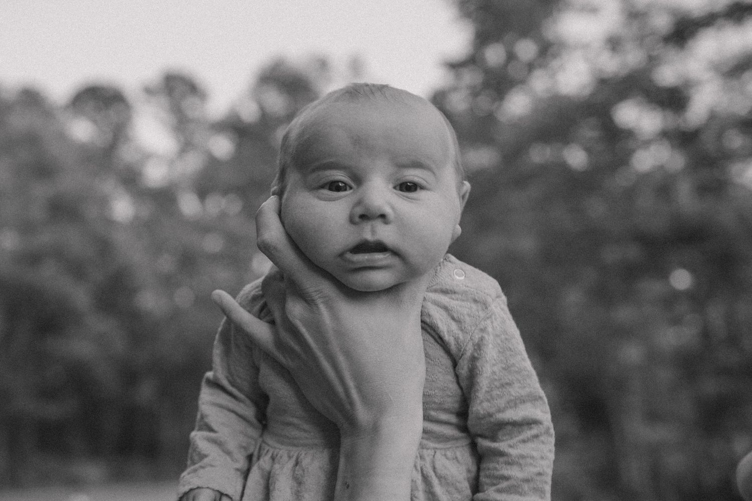 Black and white photo of a newborn baby being held up by an adult hand outdoors with trees in background.