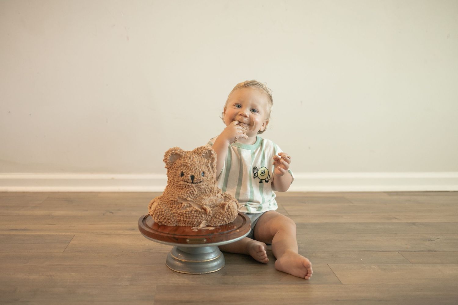 Baby smiling while eating a bear-shaped cake on a cake stand during a first birthday cake smash photo session.