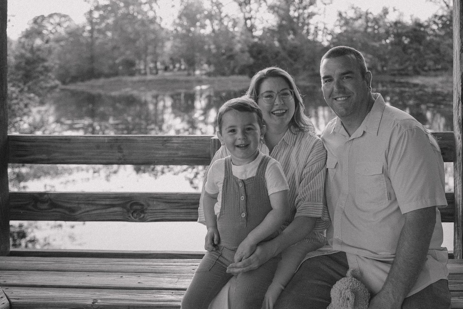 Black and white family portrait of parents and young child smiling on a bench near a scenic pond outdoors.