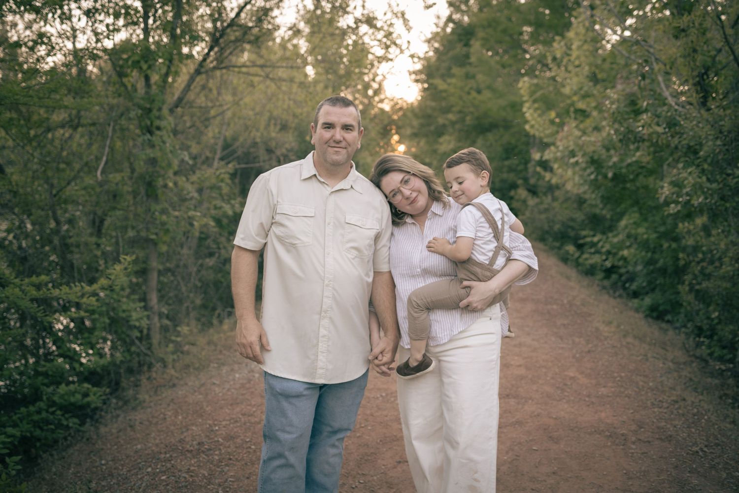 A happy family of three poses on a wooded path at sunset, with parents holding their baby boy in suspenders.