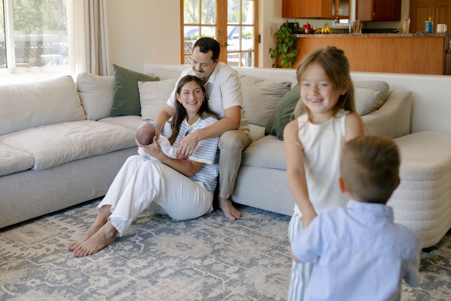 Happy family with newborn baby relaxing in bright living room while two young children play nearby.