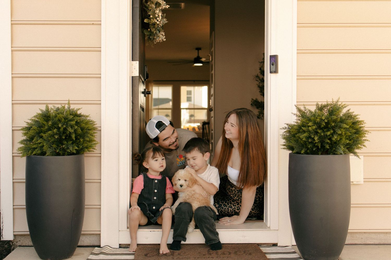 Family of four with a dog sitting on front porch steps, flanked by large planters, enjoying a warm moment together.