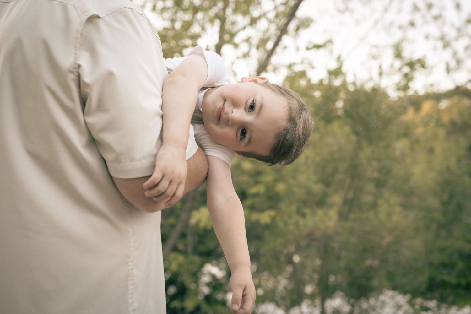 A smiling young child in a white dress being held by an adult outdoors with green trees in the background.