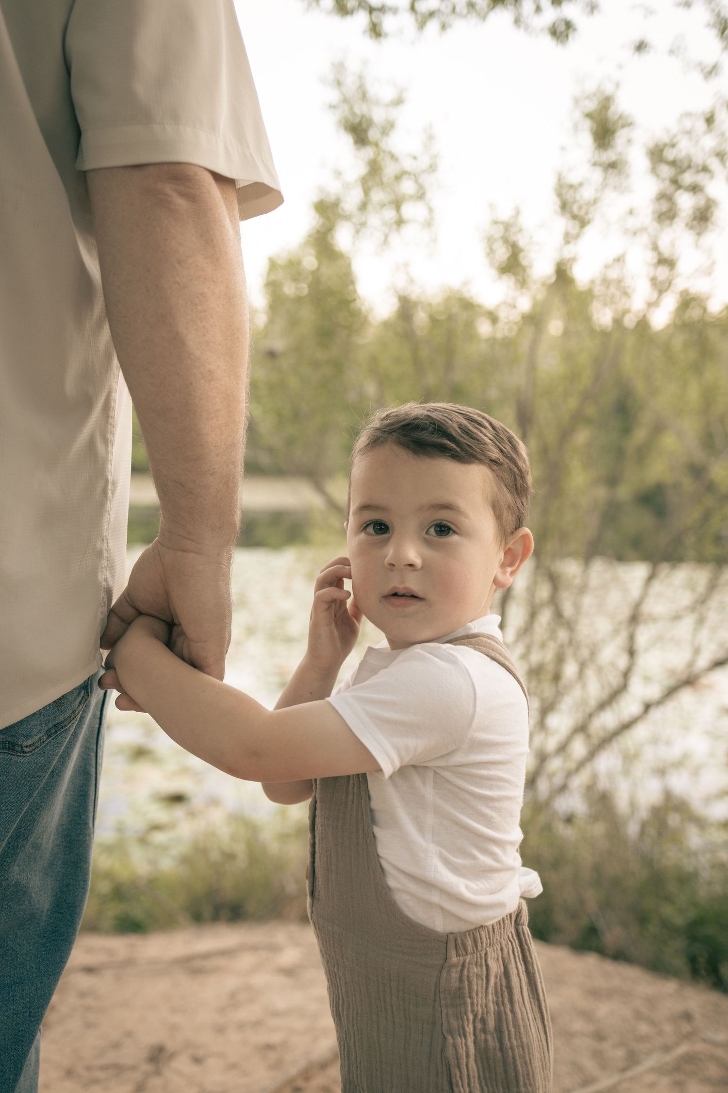A young toddler boy in white shirt and linen shorts holds his father's hand outdoors near a river.