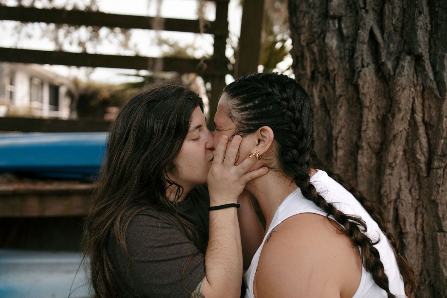 Two young women sharing a romantic kiss outdoors near a tree, captured in a candid moment.