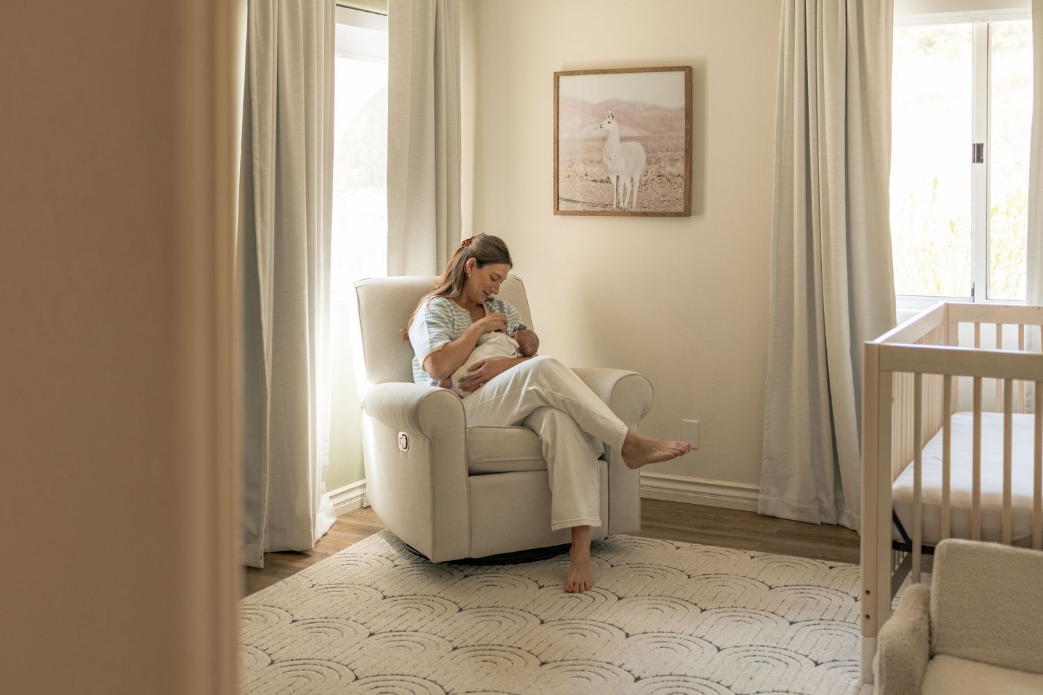 Mother sitting in white nursery glider chair cradling and bonding with newborn baby near bright window.