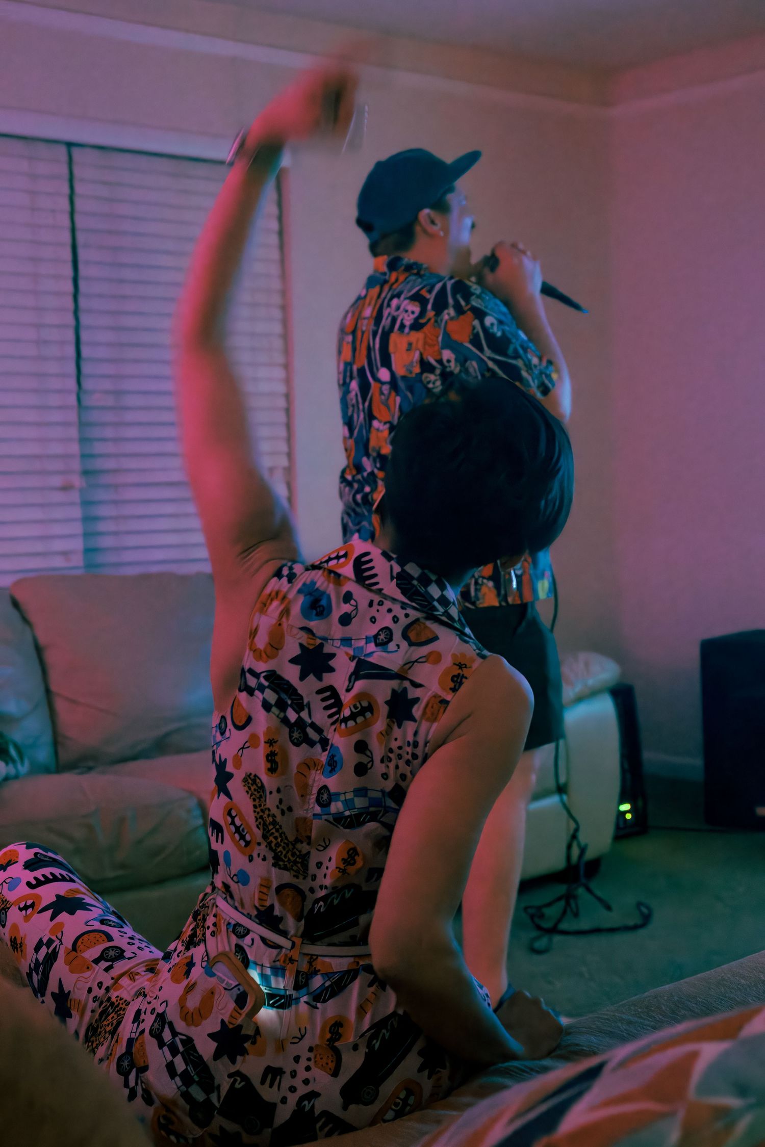 Two people in patterned outfits perform karaoke in a living room bathed in neon pink and blue lighting.