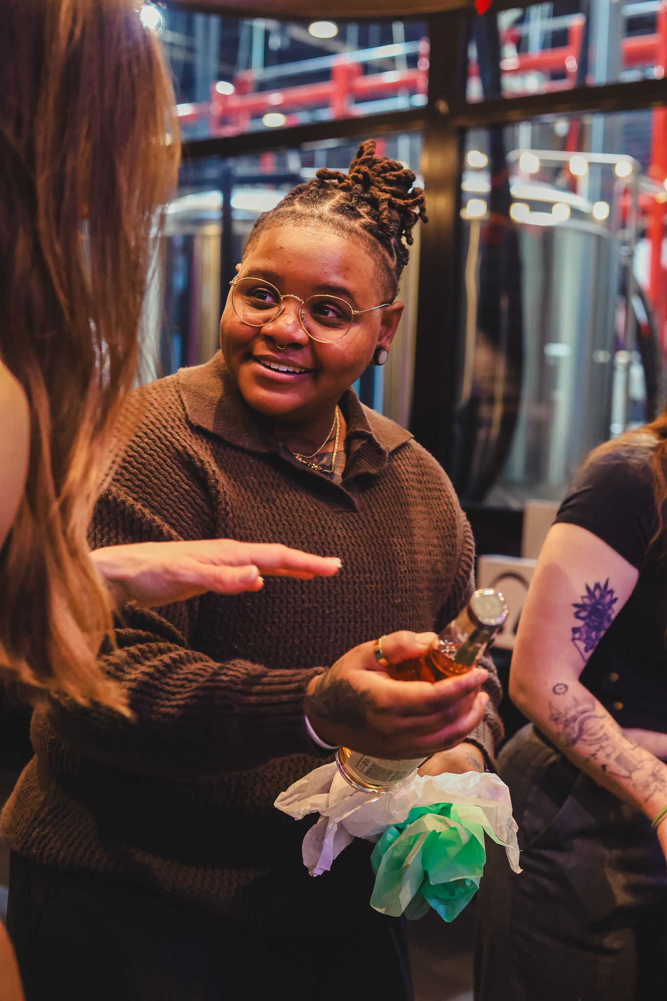 Smiling woman with braids and glasses holding bottle chatting at indoor party.