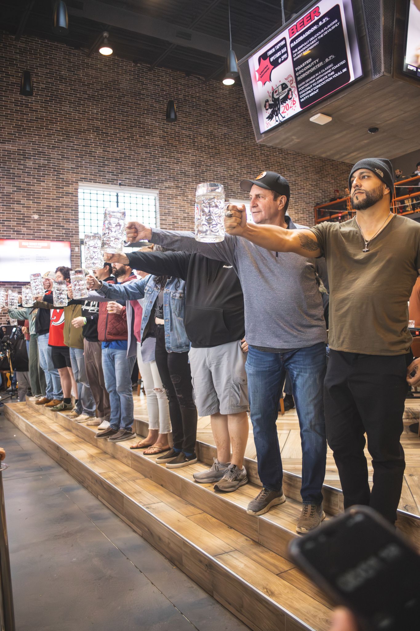 Group of people standing on a wooden platform raising glasses in a toast inside a brick-walled brewery or bar.