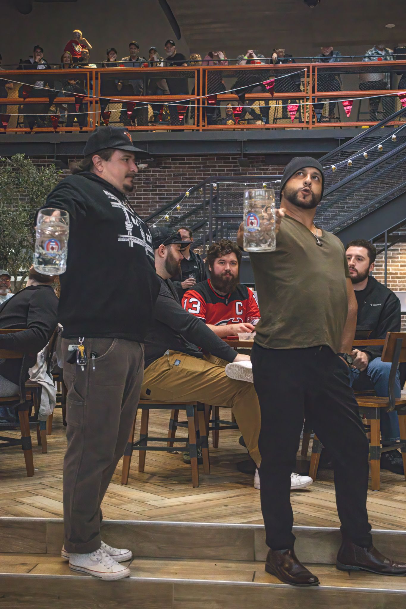Two men holding large beer steins at a lively brewery event with spectators on an upper level.