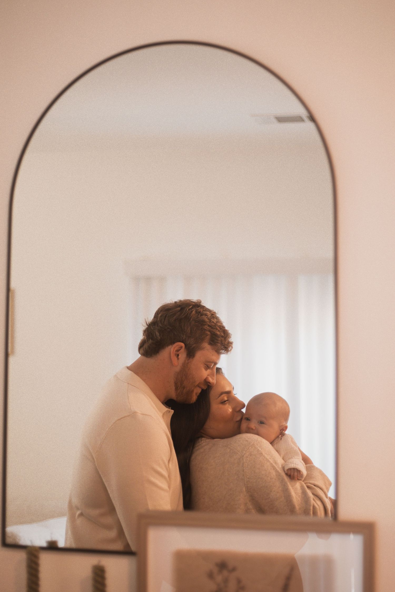 A couple tenderly holds their newborn baby, reflected in an arched mirror in a softly lit room.