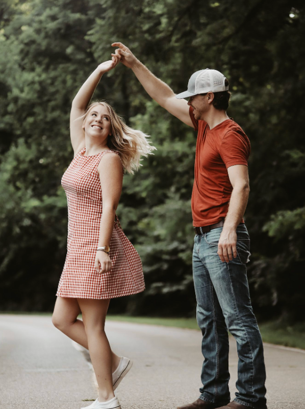 A man in a red shirt spins a laughing blonde woman in a red dress on a road.