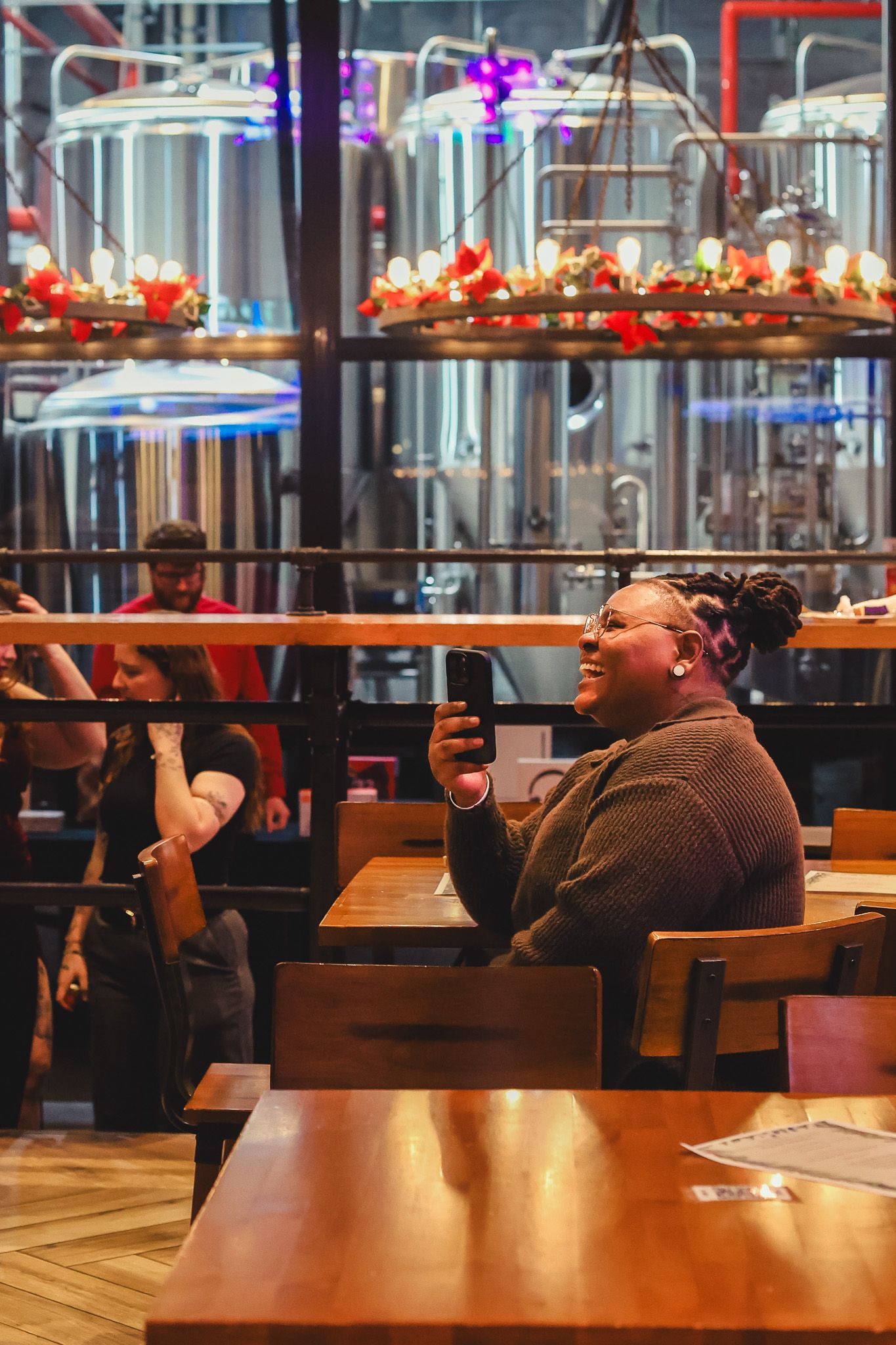 A smiling woman holds her phone at a brewery restaurant with large steel fermentation tanks in the background.