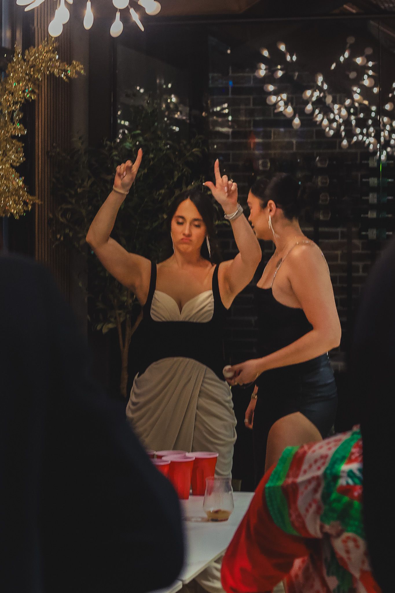 Two women dance energetically near a beer pong table at an indoor holiday party.