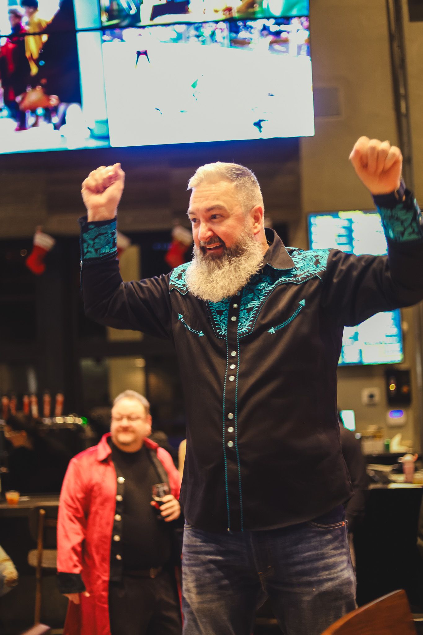 Bearded man in embroidered western shirt celebrates with arms raised at a festive bar event.