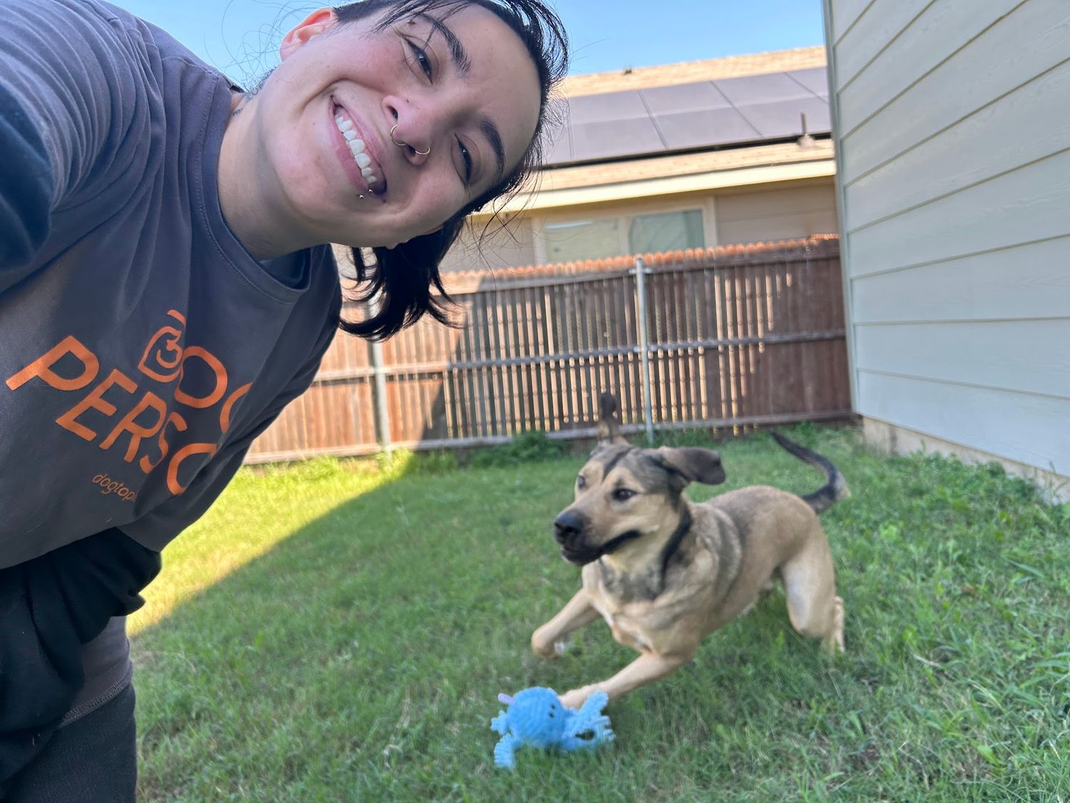 A smiling woman in a Perro shirt takes a selfie with a playful German Shepherd puppy outdoors.