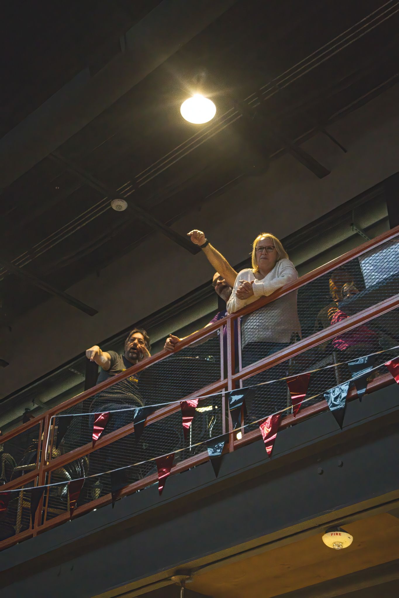 Two women cheer from a balcony railing decorated with colorful pennant flags at an indoor venue.