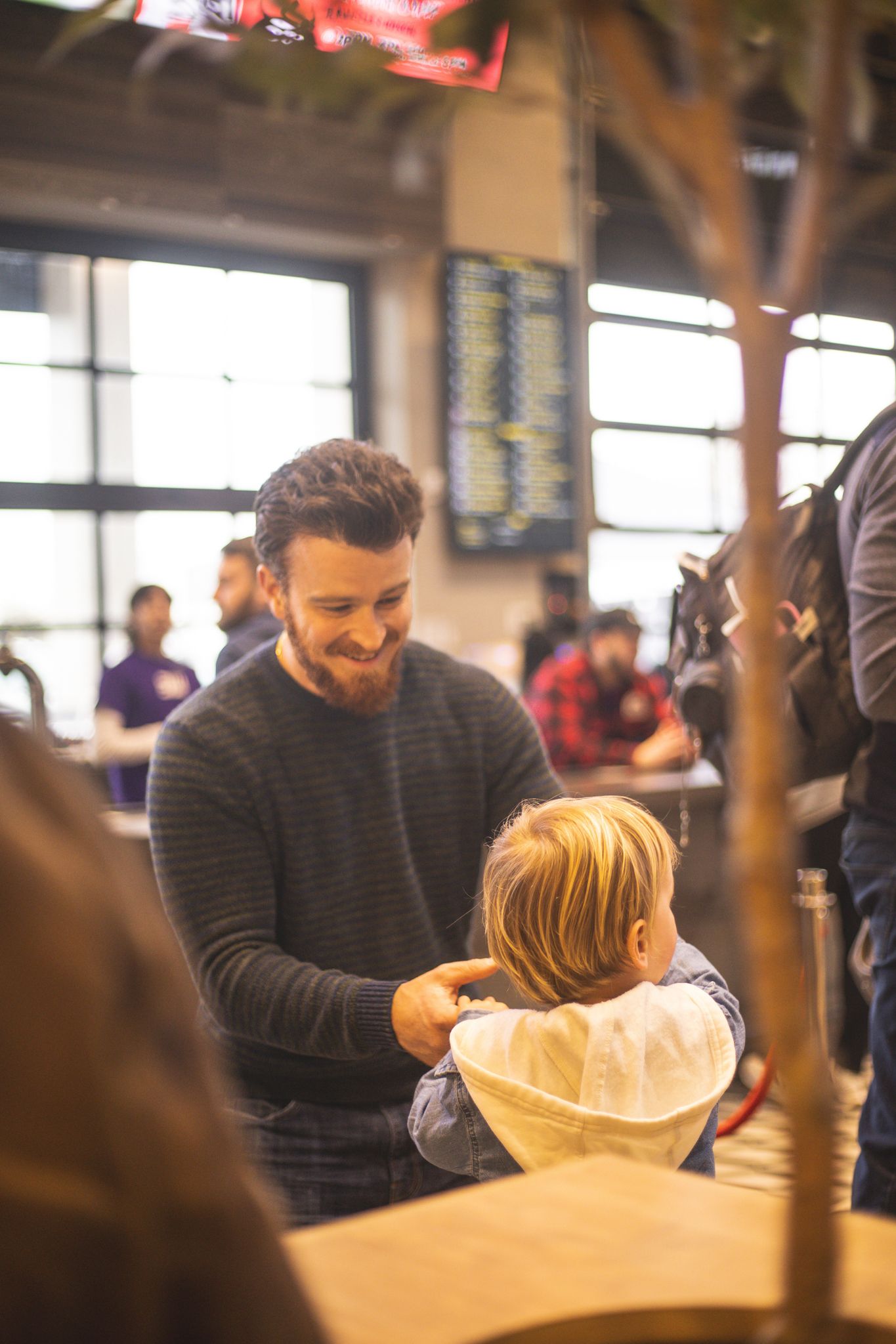 Smiling bearded man holding a young blonde child in a white hoodie inside a crowded venue.