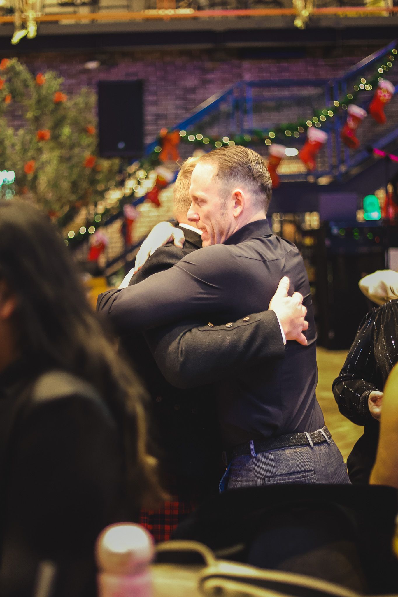 Two men share a heartfelt hug at a decorated holiday party venue with Christmas stockings in background.