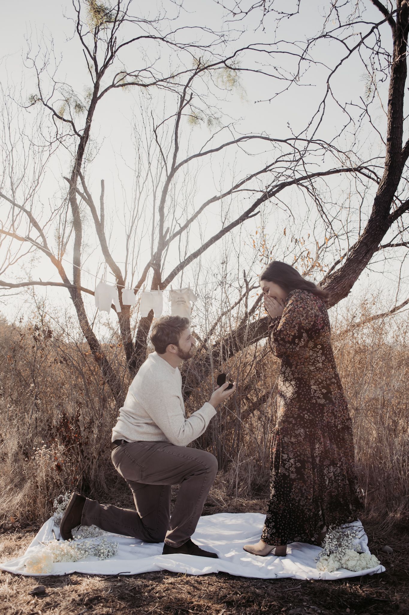 Man proposes to woman on one knee in a winter forest setting, holding a ring box while she covers her mouth in surprise.