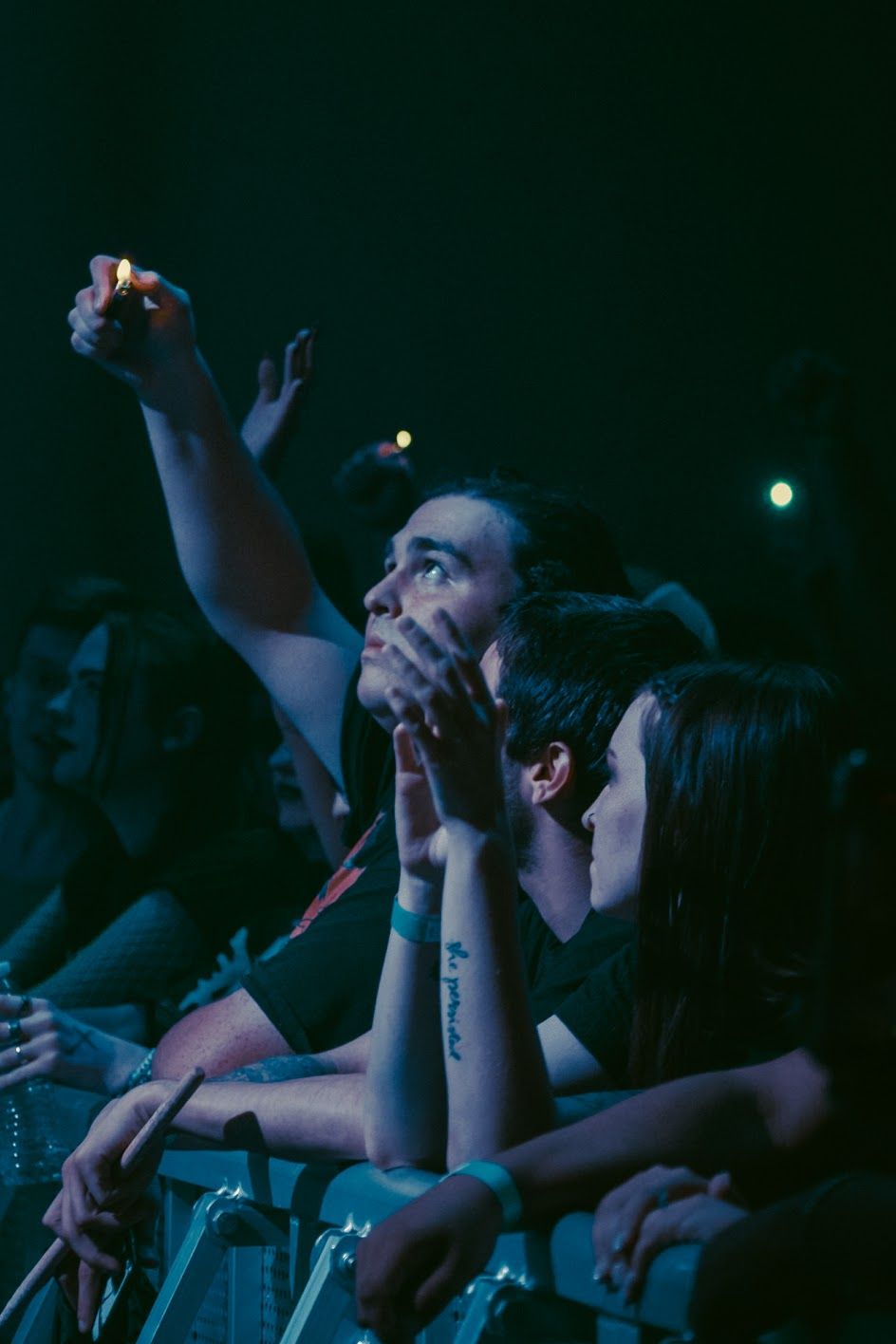 Excited concert fans in blue light, one holding up a lighter while another covers their mouth in awe.