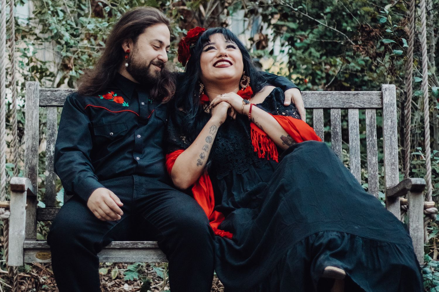 Couple in matching black and red gothic outfits sitting on a wooden bench, smiling and laughing together outdoors.