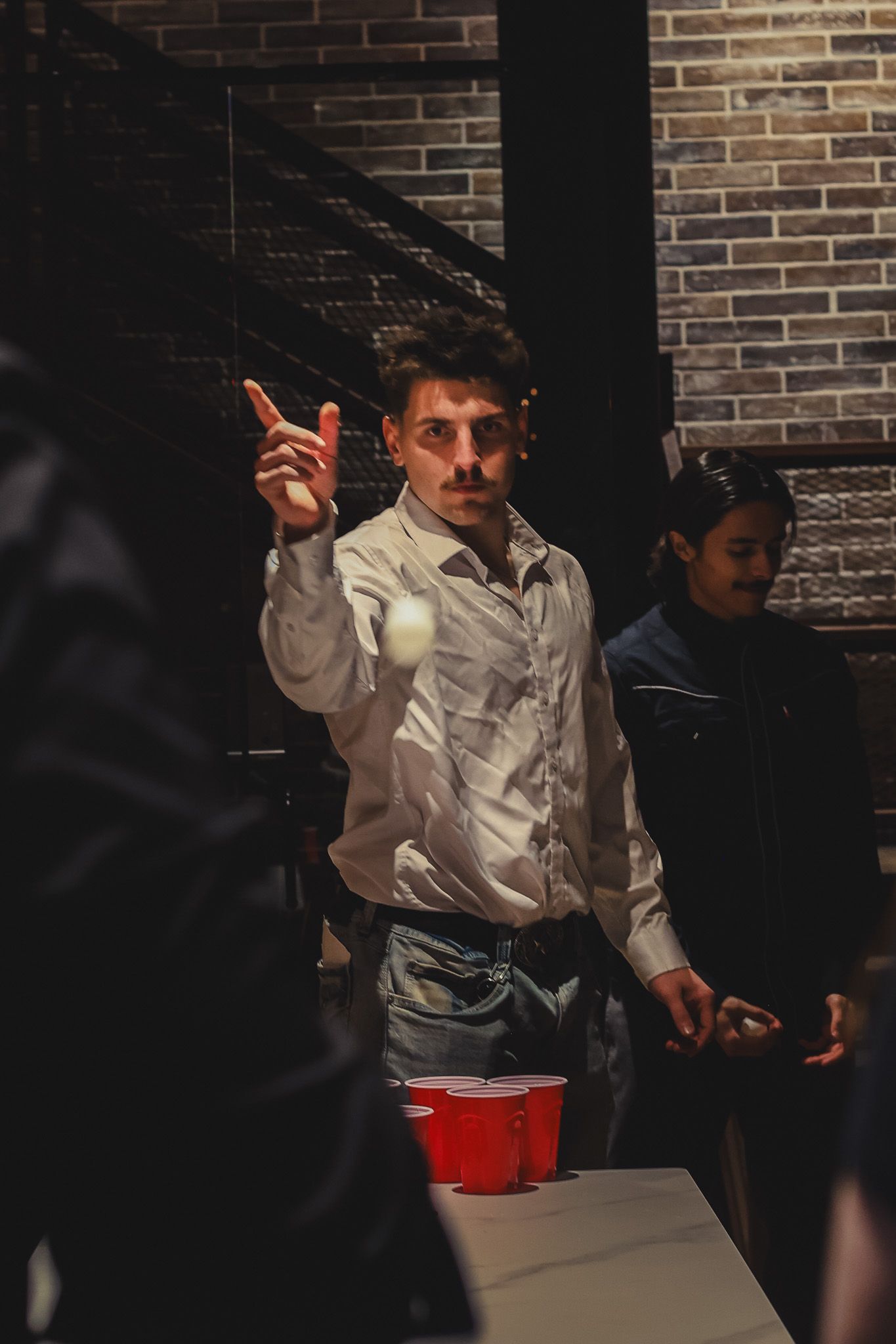 Young man tossing ping pong ball at red cups during beer pong game at a party.