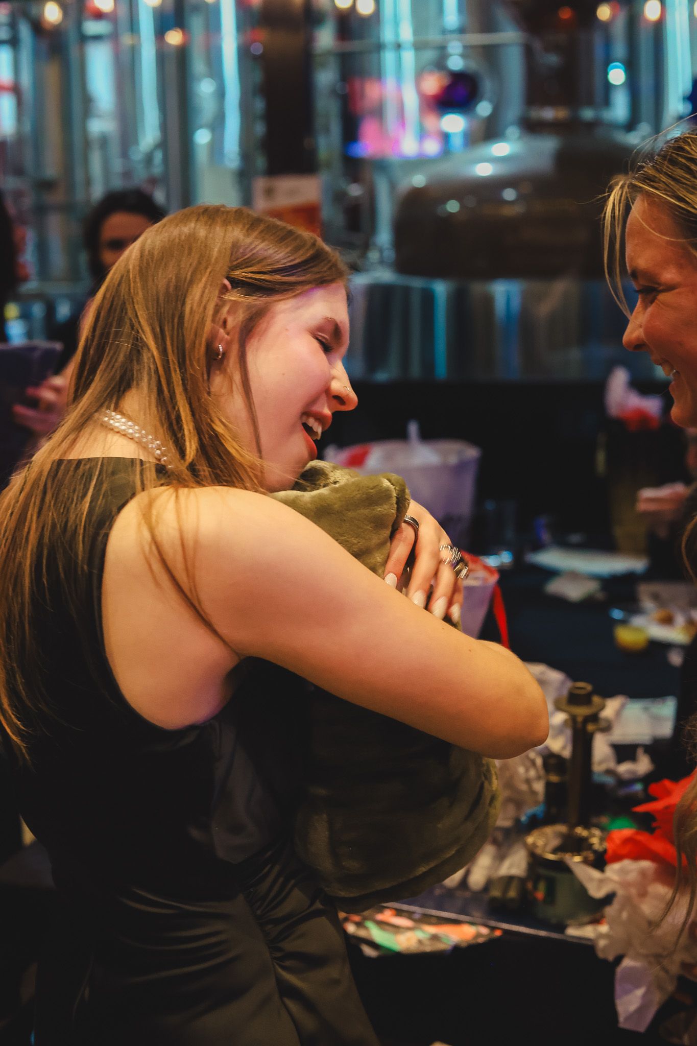 Two women sharing a warm hug and laughing together at a social event in a dimly lit venue.