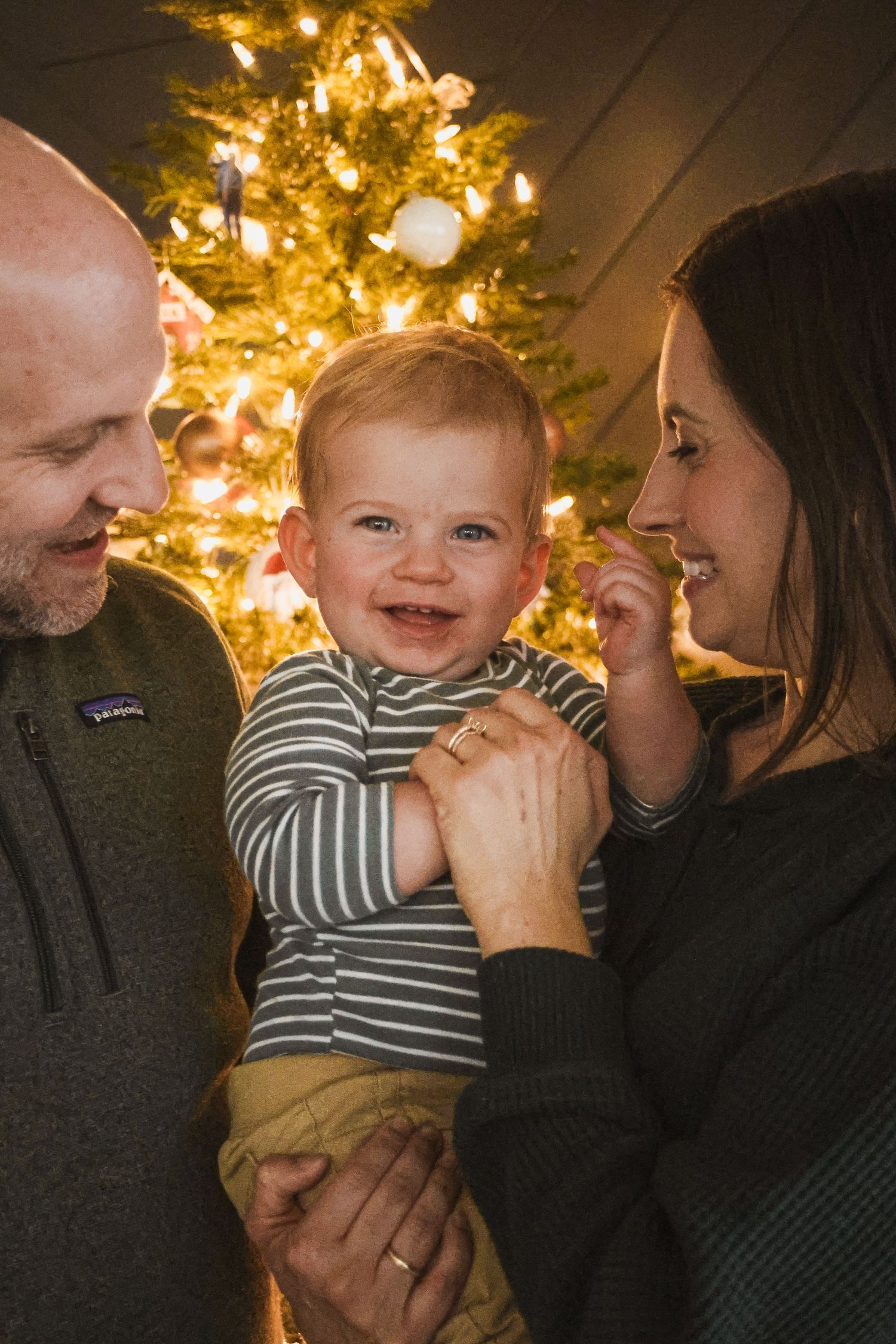 Happy baby smiles between parents in front of glowing Christmas tree during festive holiday family portrait.