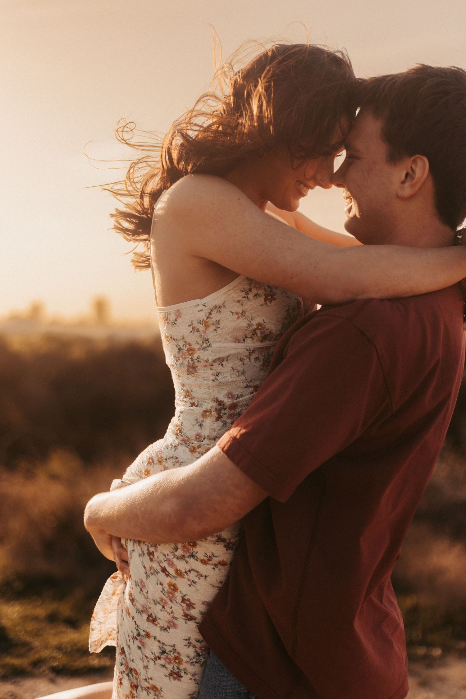 A couple embraces at golden hour, the woman in a floral dress with windswept hair leaning in close to her partner outdoors.