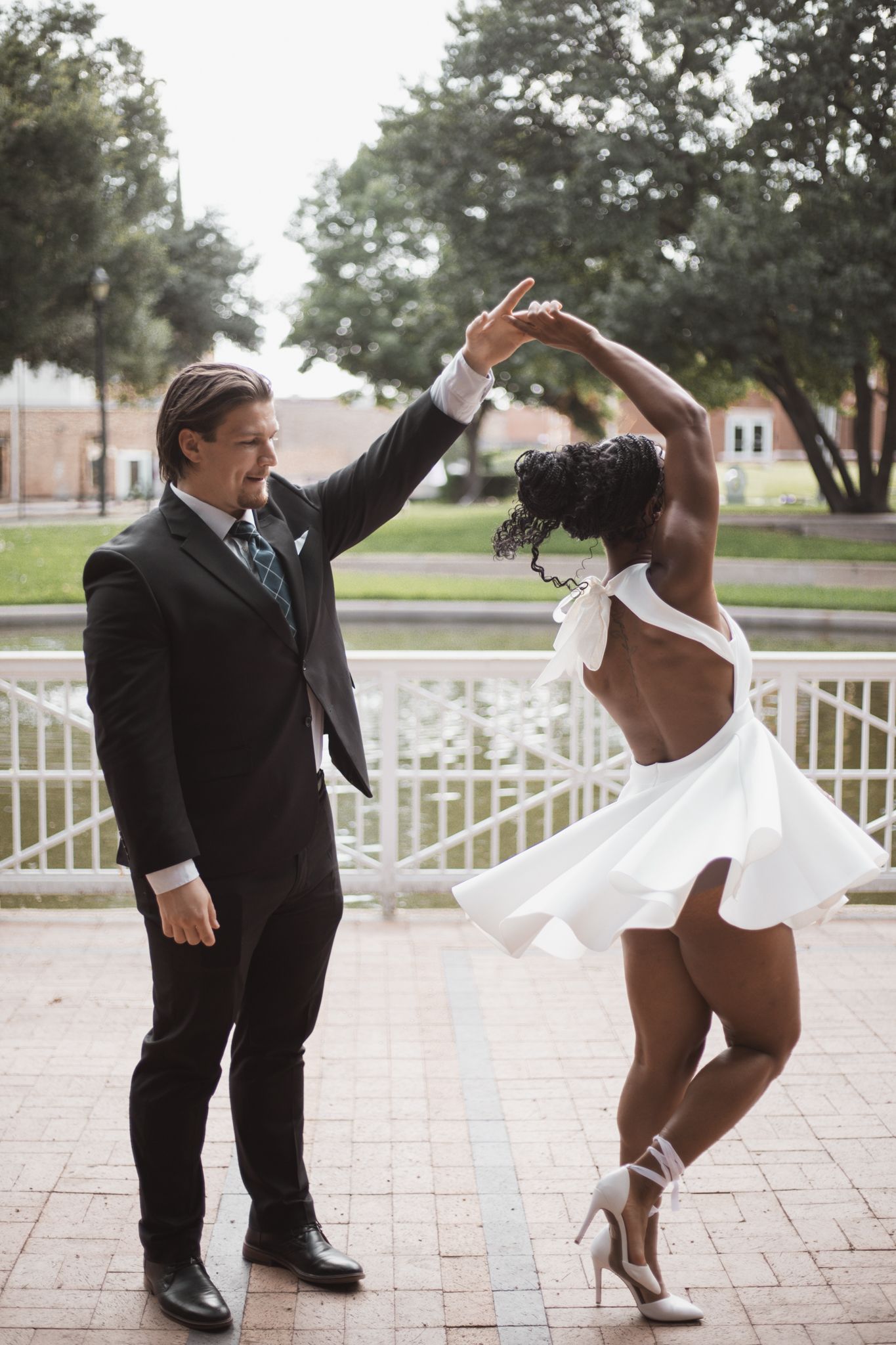 A groom in a black suit spins his bride in a short white backless dress outdoors near a fountain.