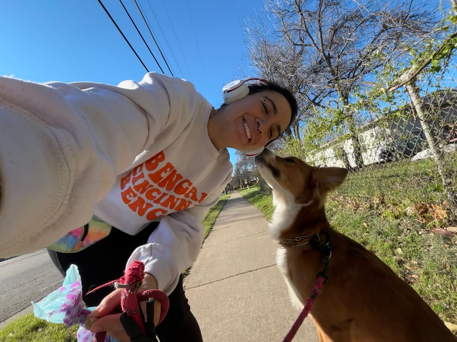 Smiling person takes outdoor selfie with a Corgi dog on a sunny tree-lined sidewalk path.