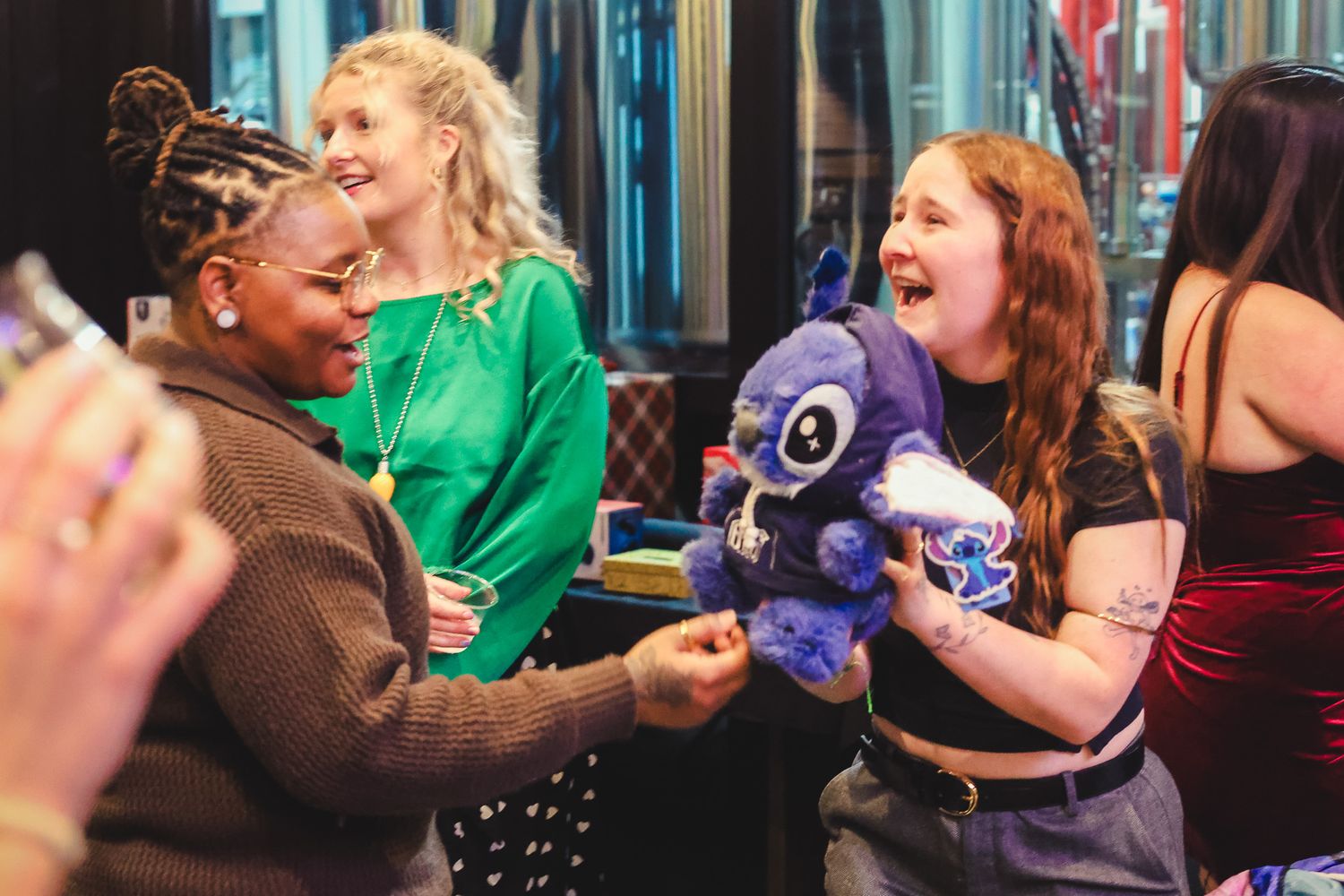 Women laughing and exchanging a blue stuffed animal toy at a lively indoor social gathering event.