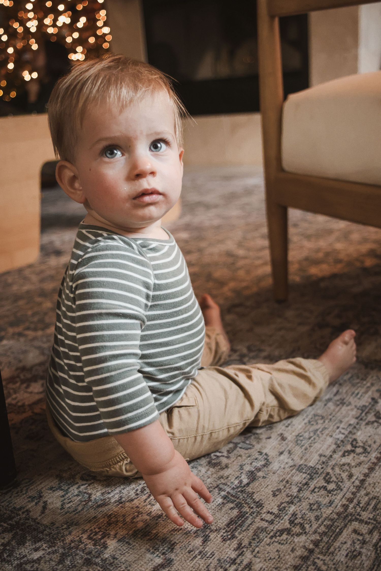 Adorable baby boy in striped shirt and khaki pants sitting on a rug near a Christmas tree.