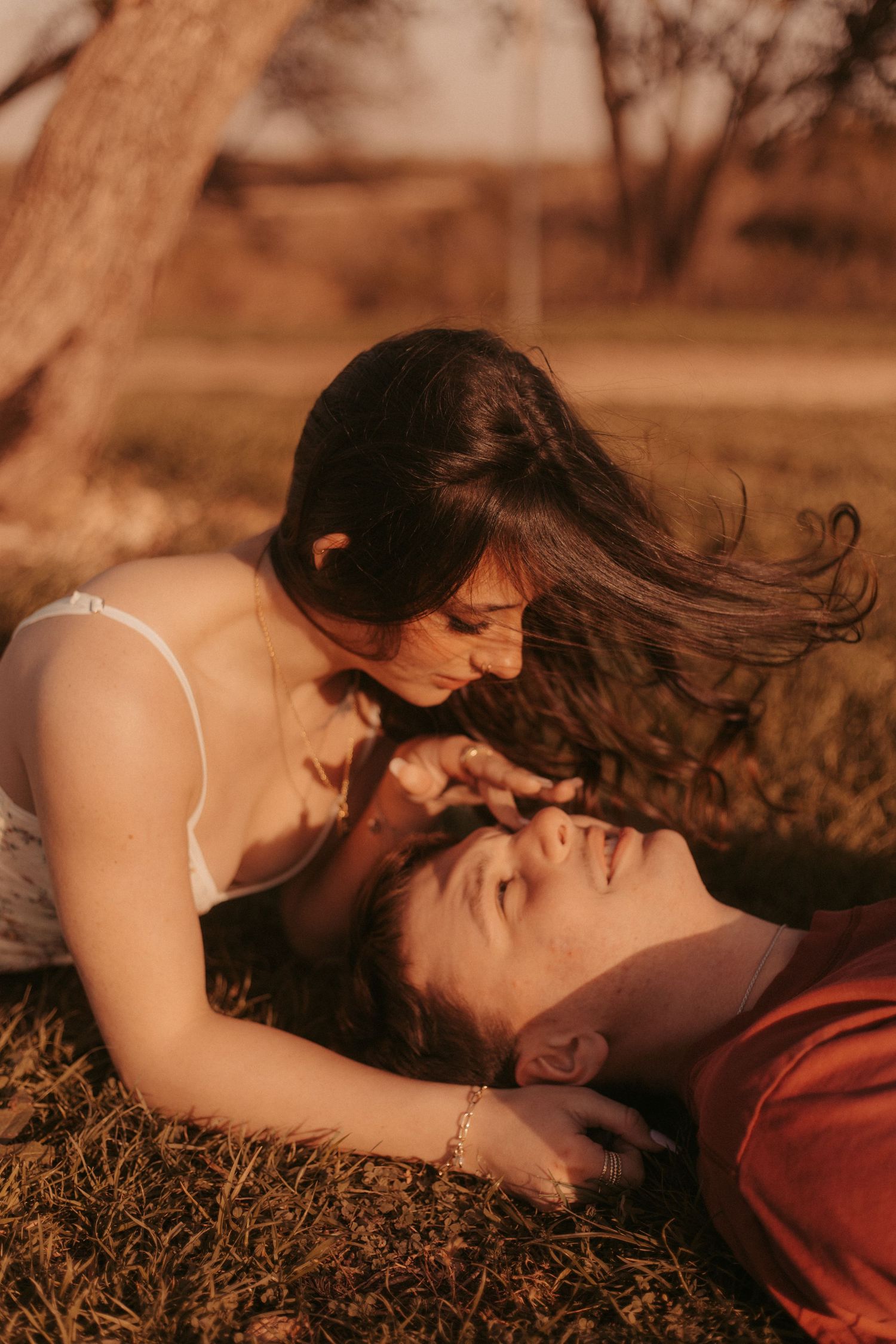 A couple shares a tender moment outdoors, woman leaning over man lying on grass during golden hour.