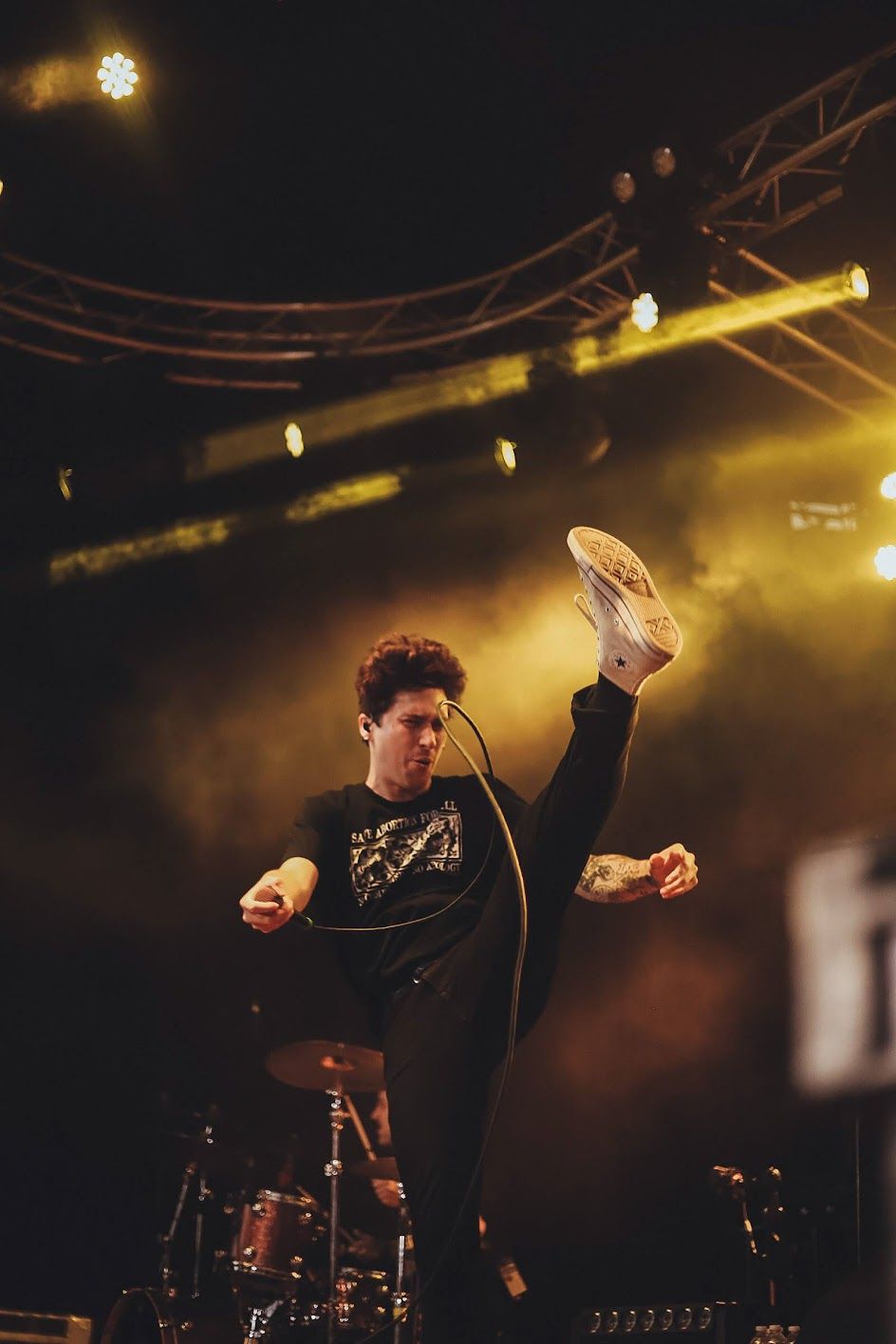 Energetic performer in black sweatshirt high-kicking on stage under dramatic yellow smoky stage lights.
